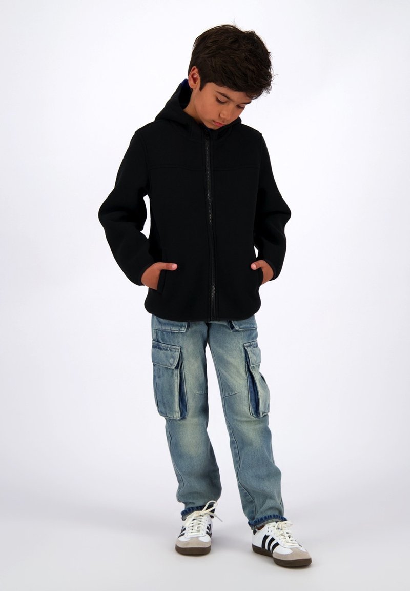 Sort zip-up hættetrøje med hætte og forlommer, parret med lyseblå cargo jeans med sidelommer og opfoldede cuffs. Hvide sneakers med sorte striber.