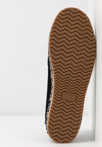 Semelle d'une chaussure TOMS noire avec un motif ondulé et le logo "TOMS" au centre de la semelle extérieure en caoutchouc marron.