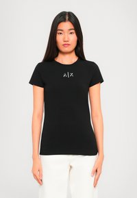 Noor naine, kellel on pikad tumedad juuksed, kannab musta lühikeste varukatega T-särki, millel on "A|X" logo, ning valgeid pükse, seistes neutraalse tausta ees.