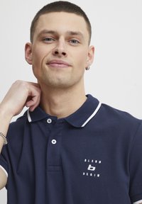 Mornarica polo majica z belimi detajli ovratnika, z dvema gumboma in vezenim logotipom "Blend Denim" na prsih. Kratki rokavi, mehak bombažen material.