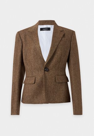 Frau trägt einen braunen Blazer über einem weißen Top, dunkelblaue gerade Jeans und schwarze offene Schuhe vor einem schlichten weißen Hintergrund.