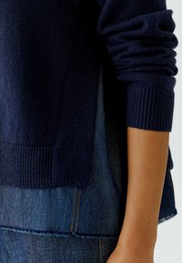Marineblau gestrickter Pullover mit gerippten Bündchen und Saum, über einem Denim-Oberteil mit ausgefransten Kanten und sichtbaren Nähdetails getragen.