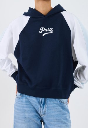 Personne portant un sweat à capuche bleu marine avec des manches blanches et le texte "Paris", associé à un jean bleu clair, mains sur les hanches.