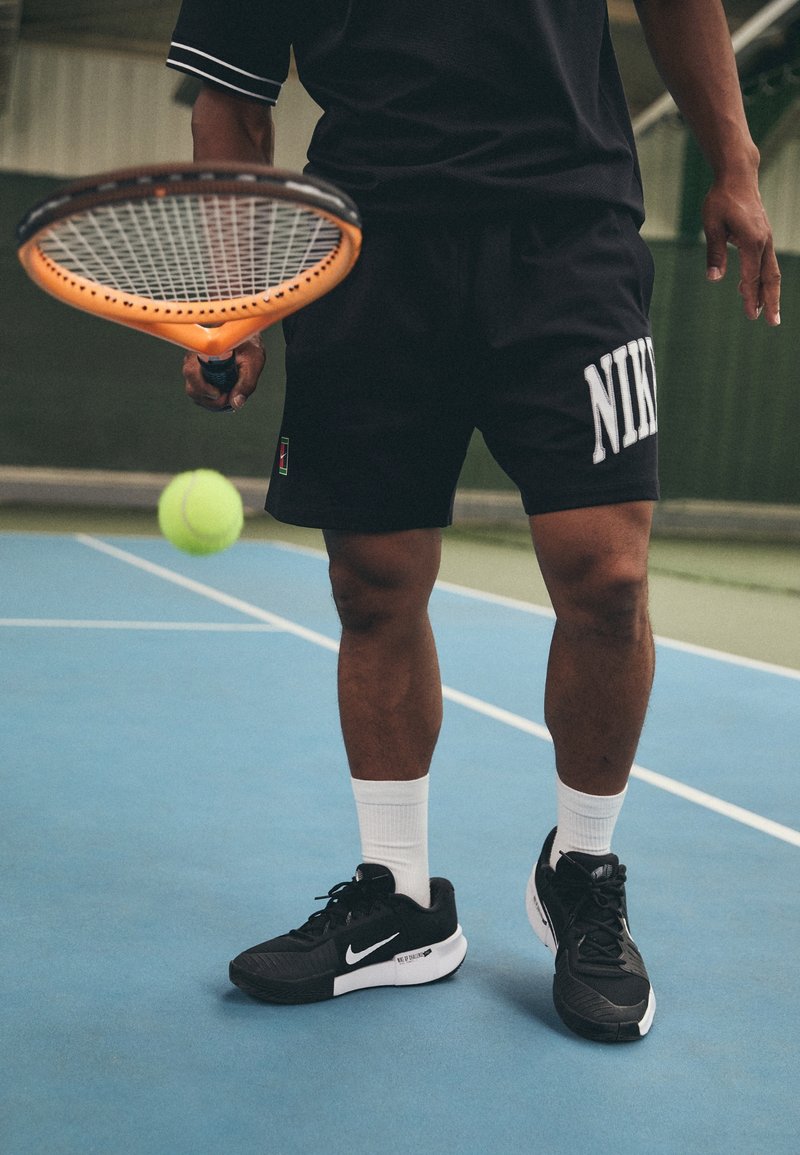 Shorts Nike noirs avec logo blanc, chaussures de sport noires, chaussettes blanches, tenant une raquette de tennis orange sur un court bleu avec un arrière-plan vert.