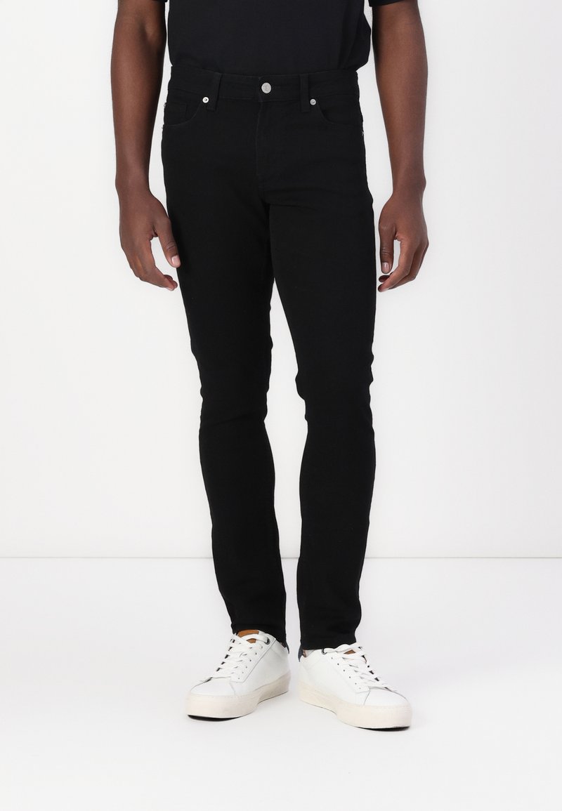 Jean denim noir, coupe slim, cinq poches, fermeture à bouton métallique, avec des détails de couture minimalistes. Associé à des baskets blanches.