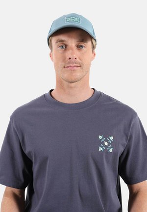 Jeune homme portant une casquette bleu clair et un t-shirt bleu foncé avec un petit motif floral bleu clair et blanc sur le côté gauche de la poitrine.
