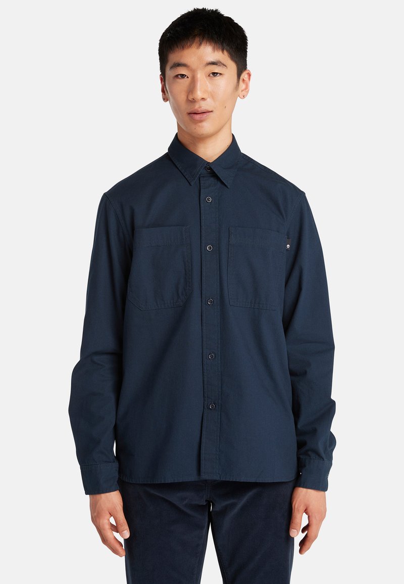 Jonge man in een donkerblauw overhemd met knopen en twee borstzakken, staand tegen een effen lichte achtergrond.
