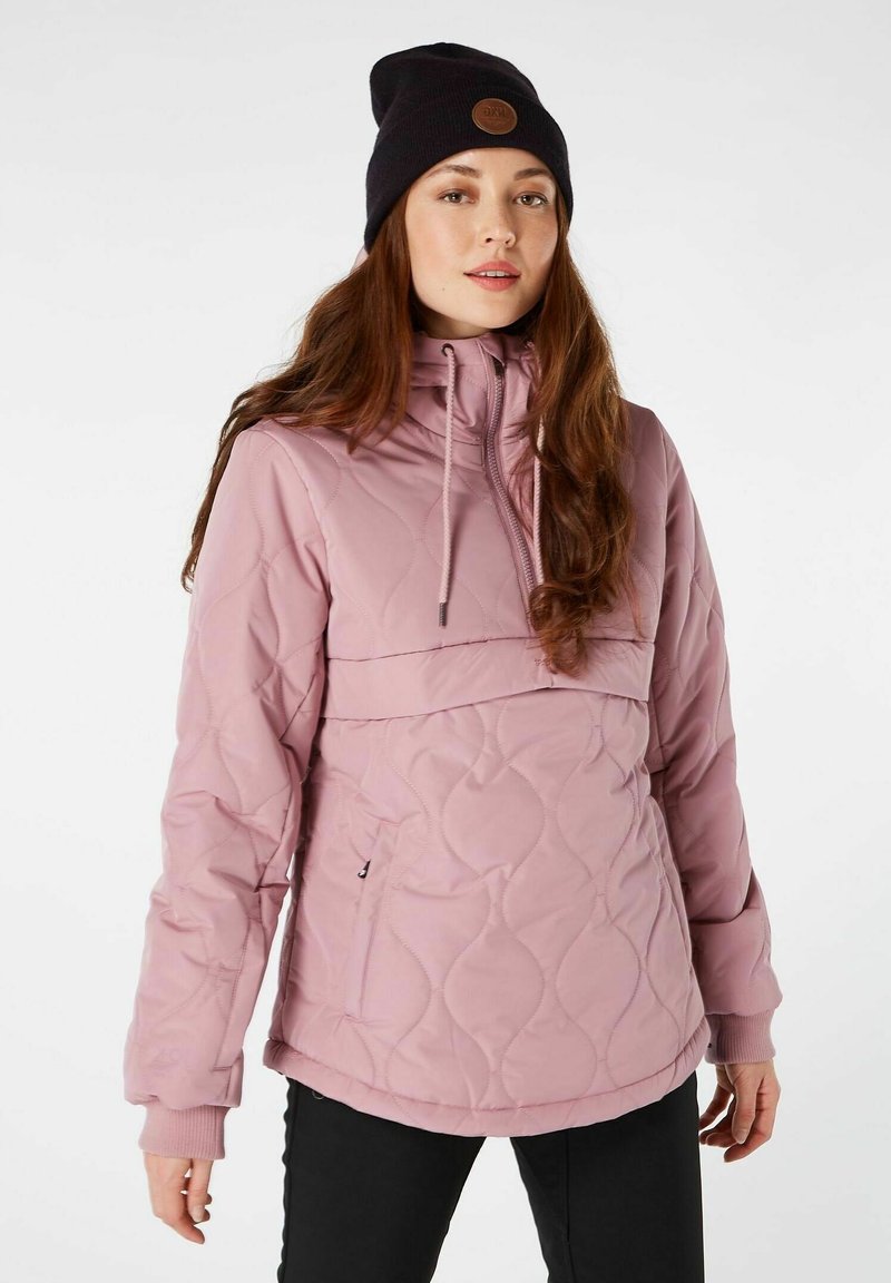 Chaqueta acolchada de color rosa claro con cuello alto, capucha con cordón y puños acanalados. Llevada sobre una blusa negra, combinada con un gorro negro.