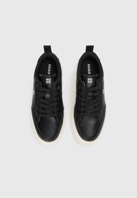 Zapatillas de cuero negro con suelas de goma crema. Las características incluyen punta redonda, diseño de cordones y logotipo grabado en la lengua.