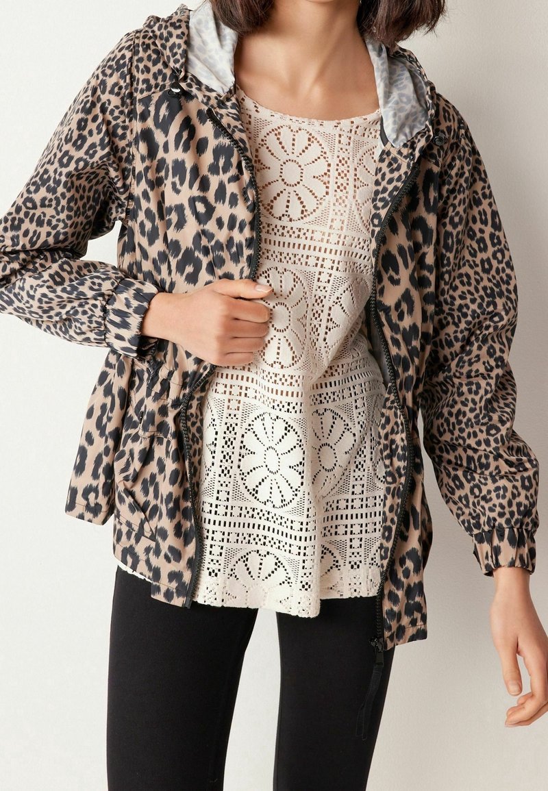 Femme portant une veste à imprimé léopard sur un haut en crochet crème et un pantalon noir, posant devant un fond clair uni.