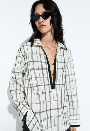 Femme aux cheveux foncés mi-longs porte des lunettes de soleil ovales noires et une chemise blanche à manches longues à carreaux avec un col en V profond et un bord noir.