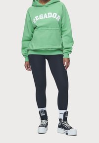 Sudadera verde con el texto "PEGADOR", bolsillo tipo canguro, combinada con leggings negros y zapatillas altas negras con suelas y detalles blancos.