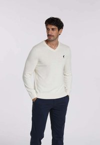 Pull en V en mélange de laine crème avec des poignets et un ourlet côtelés. Présente un petit logo noir sur la poitrine gauche. Associé à un pantalon bleu marine.