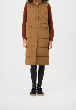 Kobieta ubrana w długą brązową pikowaną kamizelkę bez rękawów na zewnątrz rudego swetra, czarne legginsy i czarne sneakersy.