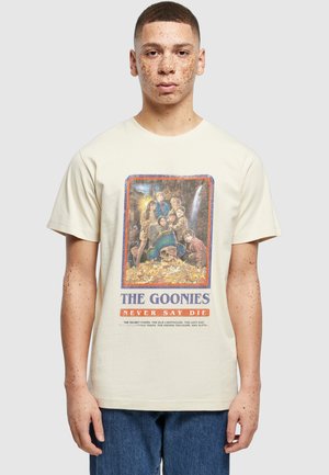 Junge Person mit kurzen Haaren und Sommersprossen, die ein beiges T-Shirt mit dem Filmplakat von The Goonies und blaue Jeans trägt.