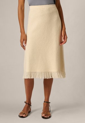 Knielanger Rock in Creme mit Fransen am Saum, getragen mit schwarzen und goldenen Riemchensandalen.