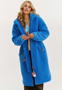 Modrá fleece bunda s kapucňou, textúrovaný povrch, zapínanie na cvoky a zips, ozdobné detaily na vreckách. Obsahuje dekoratívny prívesok na zipe.