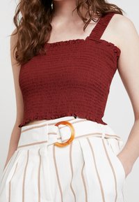 Bordeaux farvet cropped top med struktureret smocking, der har en flæseærme og en enkelt skulderstrop, parret med højlivede hvide stribede bukser med bælte.