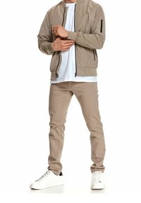Homme portant une veste bomber beige, un t-shirt blanc, un pantalon beige et des baskets blanches, ajustant la manche de sa veste avec ses mains.