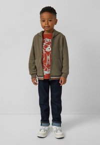 Olivengroene zip-up hoodie met twee zakken over een rode T-shirt met een beer. Donkerblauwe jeans en witte sneakers maken de outfit compleet.