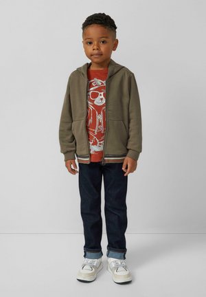 Felpa zip a oliva con due tasche sopra una maglietta rossa con grafica di un orso. I jeans blu scuro e le sneakers bianche completano il look.