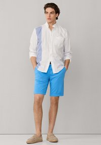 Uomo in piedi con le mani in tasca, indossa una camicia bianca con bottoni e righe blu e pantaloncini blu vivaci su uno sfondo semplice.