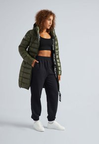 Groene pufferjack met horizontale stiksels, zwarte crop top, zwarte joggingbroek en witte sneakers. De jas heeft logo-accenten.