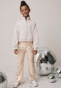 Felpa crema con zip e spalle scese, abbinata a pantaloni svasati con paillettes dorate. Scarpe da ginnastica bianche completano il look, con sfere da discoteca specchiate sullo sfondo.