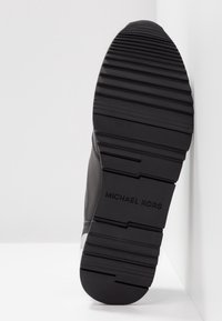 Semelle d'une chaussure noire Michael Kors avec des rainures horizontales profondes, inclinée sur fond blanc montrant un motif de la bande de roulement texturé.