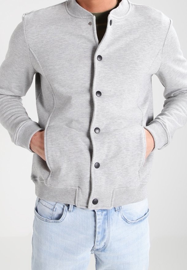 Homme portant une veste bomber gris clair boutonnée, les mains dans les poches et un jean bleu clair sur un fond uni.
