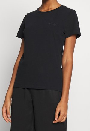 Personne portant un t-shirt noir ajusté à manches courtes avec un logo brodé subtil sur la poitrine et un pantalon noir ample.