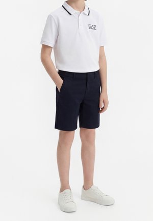 Barn iført en hvid polo med EA7-logo, marineblå shorts, hvide sneakers, stående med den ene hånd i lommen på hvid baggrund.