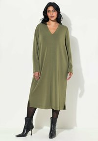 Abito lungo verde olive a maniche lunghe con scollo a V, spacchi laterali e vestibilità rilassata, abbinato a stivali neri alla caviglia, su uno sfondo neutro.