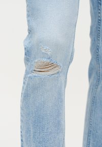 Jasnoniebieskie dżinsy z denimu z postrzępionym, poziomym rozcięciem odsłaniającym nitki i skórę w okolicy kolana.