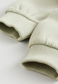 Detail béžových rukávů mikiny s žebrovanými elastickými manžetami, ležících na světlém povrchu.