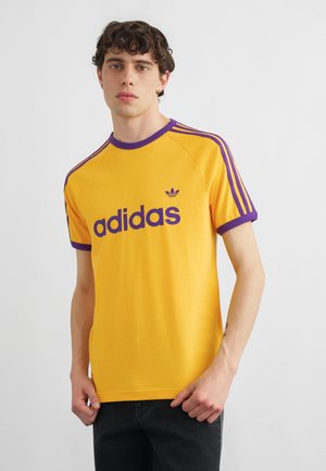 Mladý muž s kudrnatými tmavými vlasy, oblečený v žlutém tričku Adidas s fialovými detaily a černých kalhotách na jednoduchém pozadí.