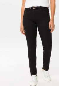 Jeans skinny neri con vita media, passanti per cintura e dettagli in oro, abbinati a sneakers bianche su uno sfondo neutro.