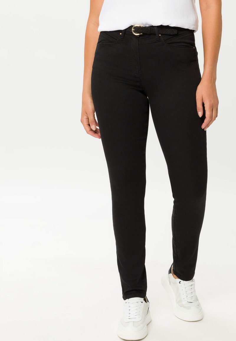 Jean skinny noirs avec une taille mi-haute, passants de ceinture et accents en matériel doré, associés à des baskets blanches sur un fond uni.