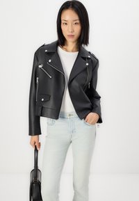 Veste de moto en simili cuir noir avec des zippers argentés et des boutons-pression, coupe cropped, portée sur un top blanc et associée à un jean bleu clair.