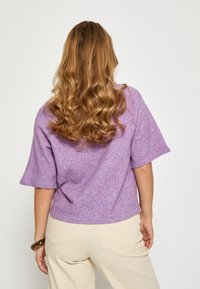 Violeta, brīvi pieguļošs, īso piedurkņu džemperis ar teksturētu adījumu. Iezīmējal casual stilu, noapaļots apakšmala, un tas ir apvienots ar platām, bēšām biksēm.