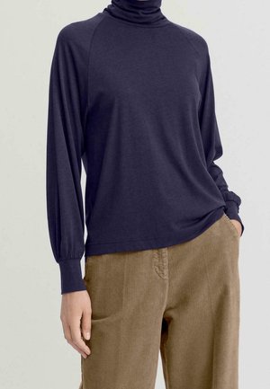 Mujer con top de cuello alto azul marino de manga larga y pantalones de pana marrón claro, con una mano en el bolsillo, contra un fondo liso.