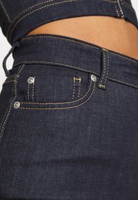 Jeans de mezclilla oscura con costuras amarillas, que cuentan con un bolsillo frontal y una textura suave. La cinturilla tiene un detalle de diseño sutil.