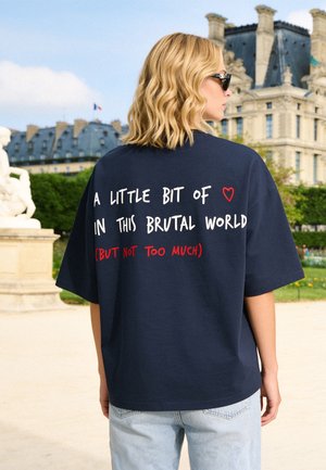 Personne blonde portant des lunettes de soleil et une chemise bleu marine avec le texte "A little bit of heart in this brutal world (but not too much)" à l'extérieur près de bâtiments historiques.