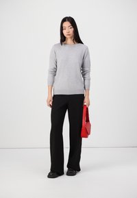 Grauer Strickpullover mit geripptem Saum, kombiniert mit schwarzen weiten Hosen. Hält eine rote Handtasche. Schwarze Schuhe mit dicker Sohle.