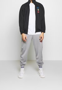 Muž v černé zipové bundě s třemi barevnými logy Adidas, bílé tričko, šedé tepláky Adidas, pruhované ponožky a bílé tenisky.