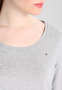 Frau trägt ein hellgraues Rundhals-Shirt mit einem kleinen roten, weißen und blauen Tommy Hilfiger-Logo auf der Brust, die Lippen leicht zusammengepresst.