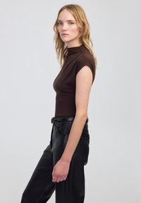 Top aderente marrone con colletto alto e maniche corte a cappuccio, abbinato a pantaloni in pelle nera a vita alta. Tessuto liscio, design semplice.