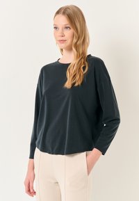 Maglietta nera a maniche lunghe con scollatura rotonda realizzata in materiale morbido; design leggermente cropped con vestibilità rilassata, abbinata a pantaloni di colore chiaro.