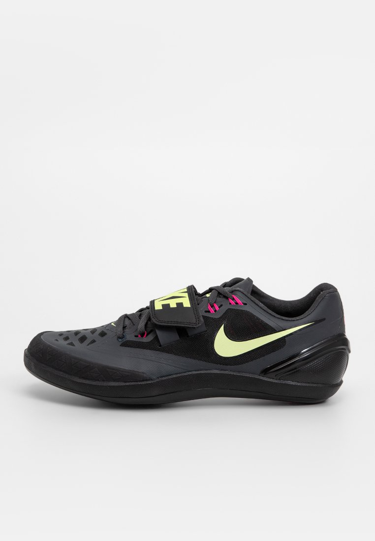 Svart Nike träningsskor med gul swoosh, rosa snöröglor och ett gult "NIKE"-band över snörningen, visad från sidan på en vit bakgrund.