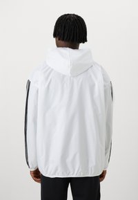 Casaco de vento branco com capuz, apresentando riscas verticais pretas ao longo das mangas e bainha elástica para um ajuste justo.